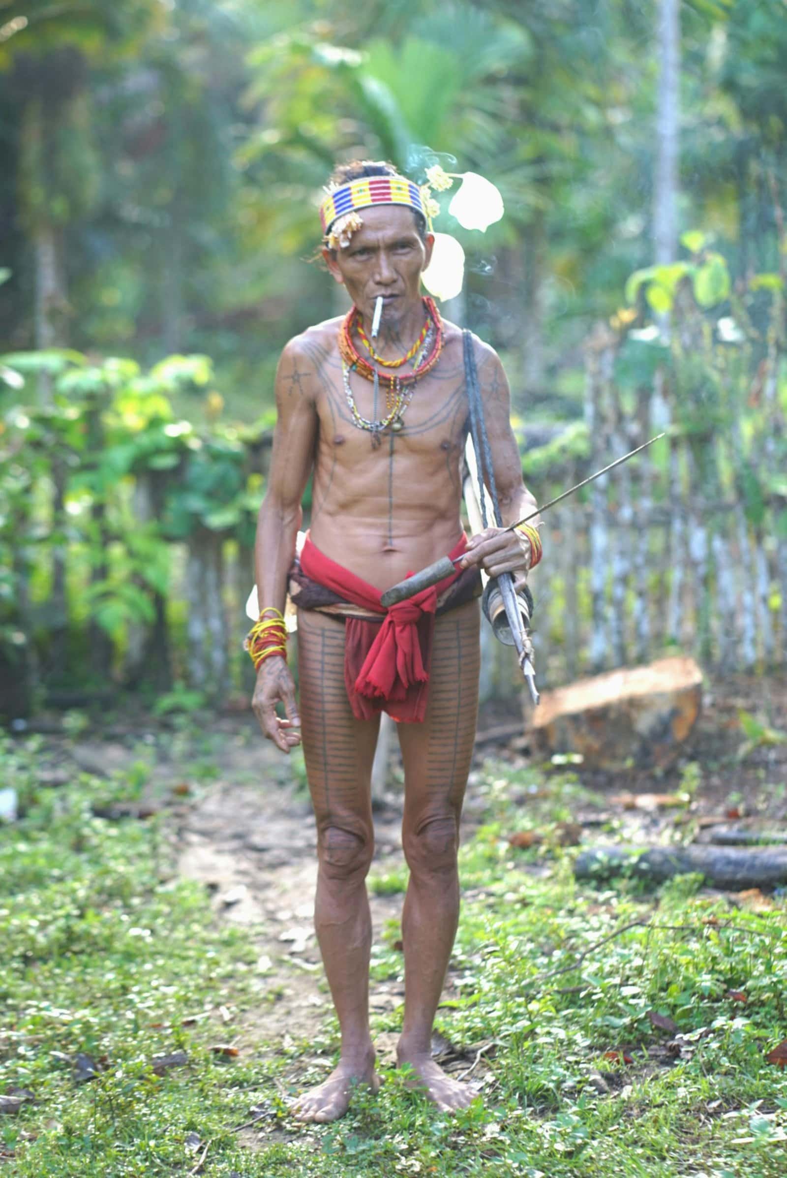 Shirtless tribal warrior with tattoos and bow in West Sumatra forest setting.